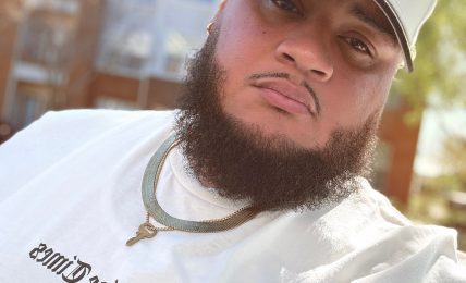 Young man with a beard wearing a white cap and white shirt outdoors, with buildings in the background.