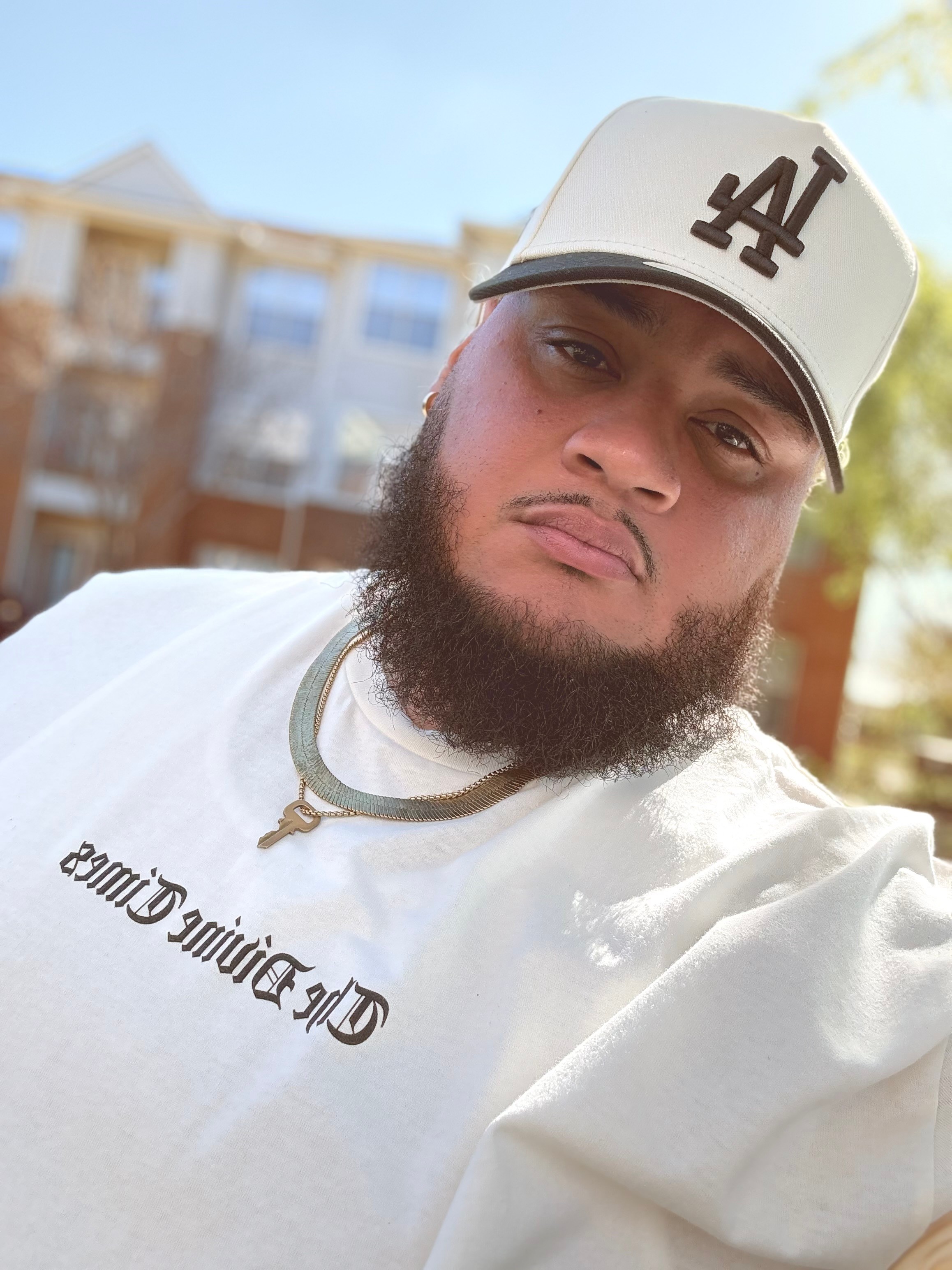 Young man with a beard wearing a white cap and white shirt outdoors, with buildings in the background.