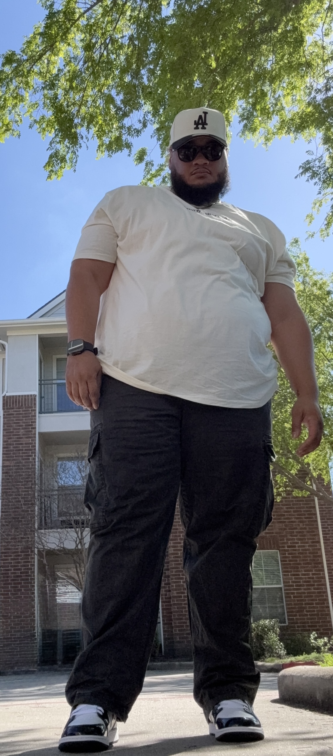 Person standing outdoors under trees, wearing a white cap, sunglasses, white t-shirt, black pants, and sneakers.