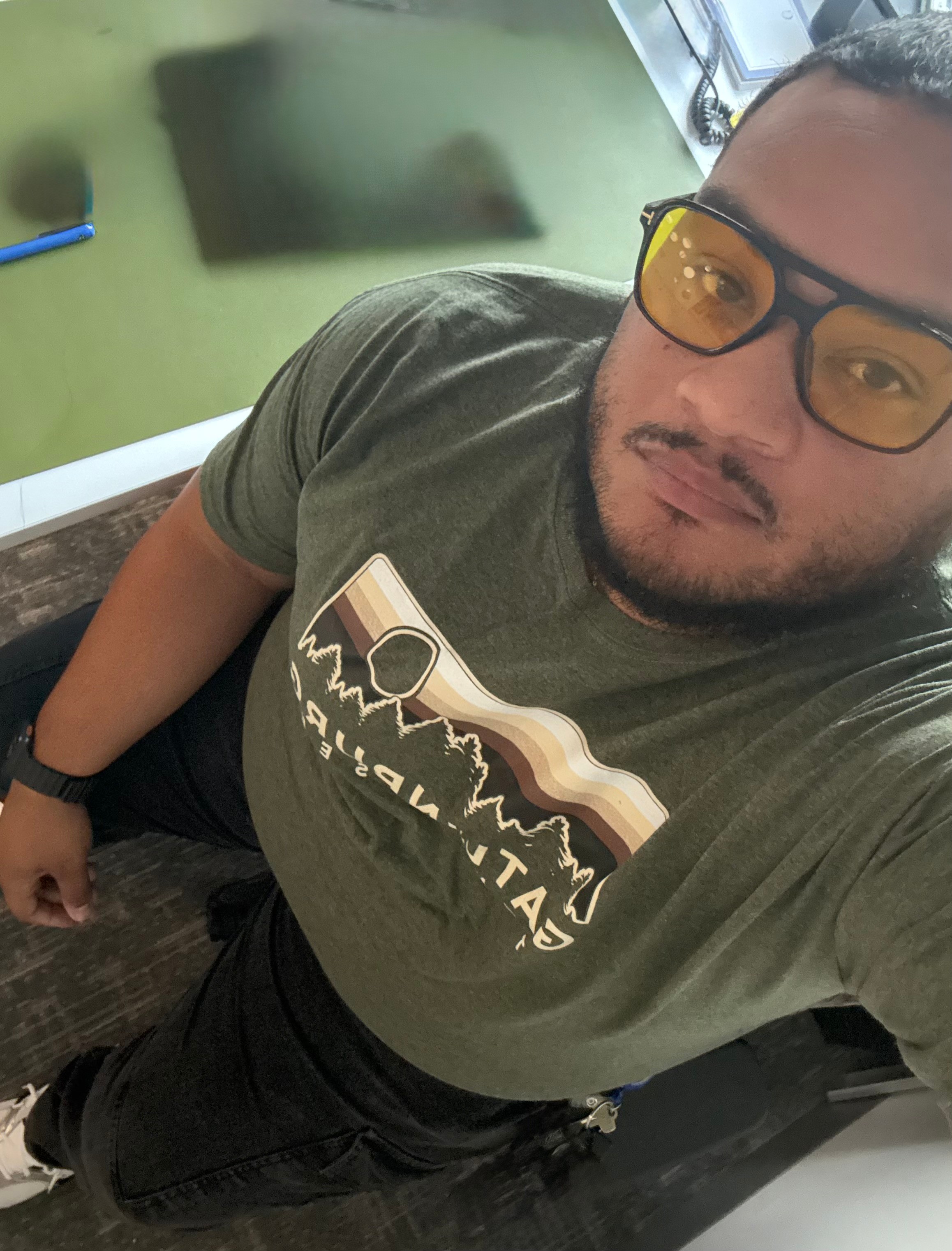 Person wearing glasses and a green T-shirt with a mountain logo, taking a selfie indoors. Green wall and window in background.