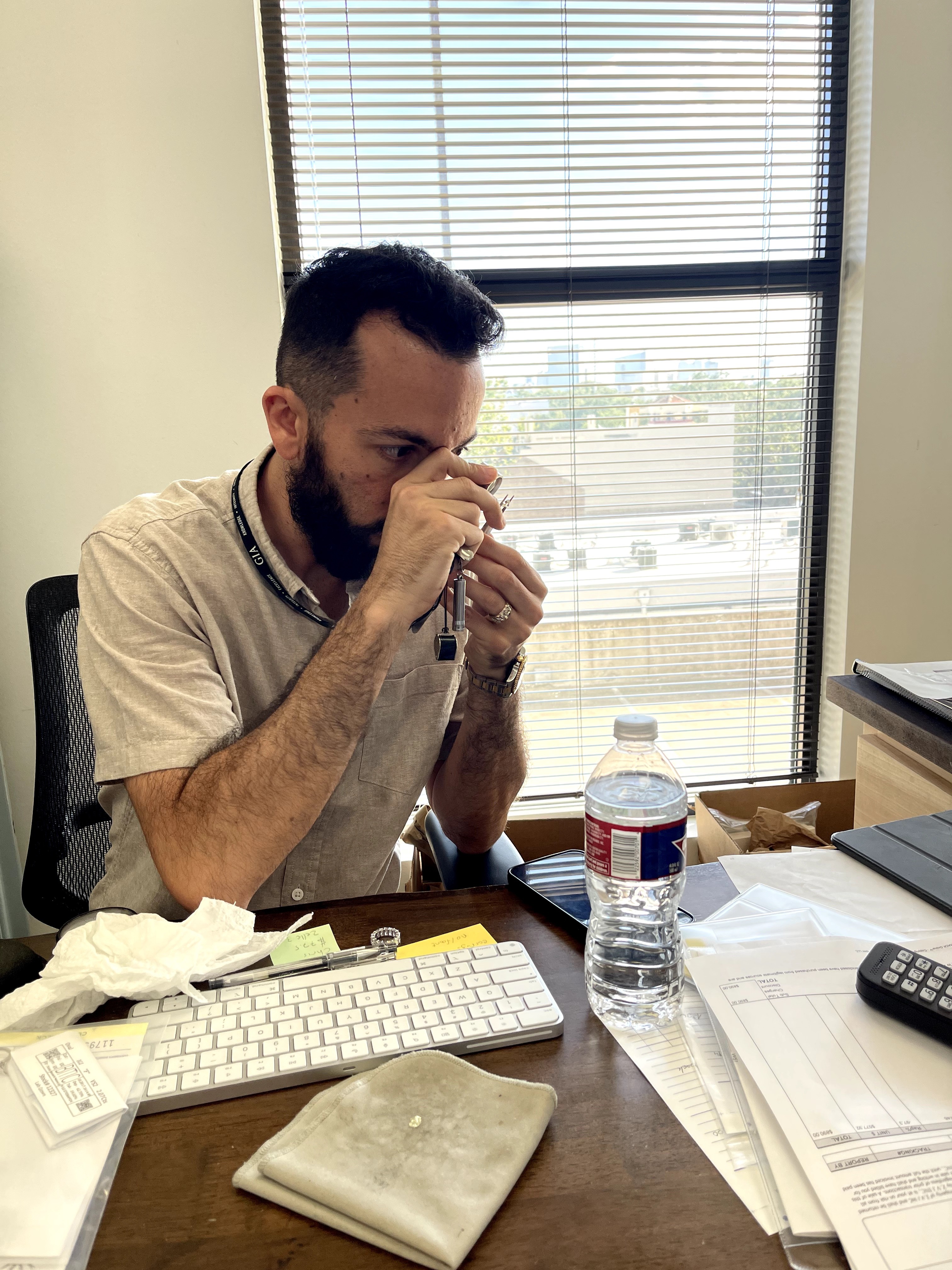 Man sitting at desk, drinking from a cup, with a window behind him showing a cityscape, office supplies on desk.