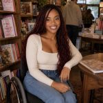 Smiling woman with long curly hair sitting in a wheelchair inside a bookstore, surrounded by shelves of books.