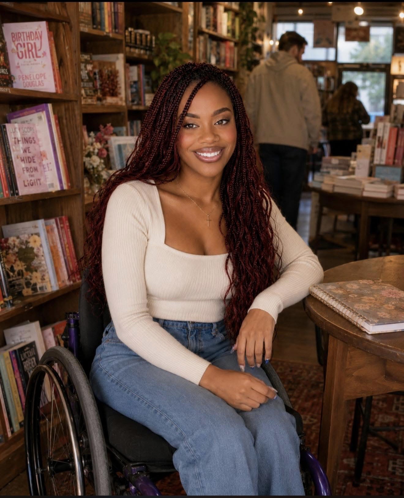Smiling woman with long curly hair sitting in a wheelchair inside a bookstore, surrounded by shelves of books.