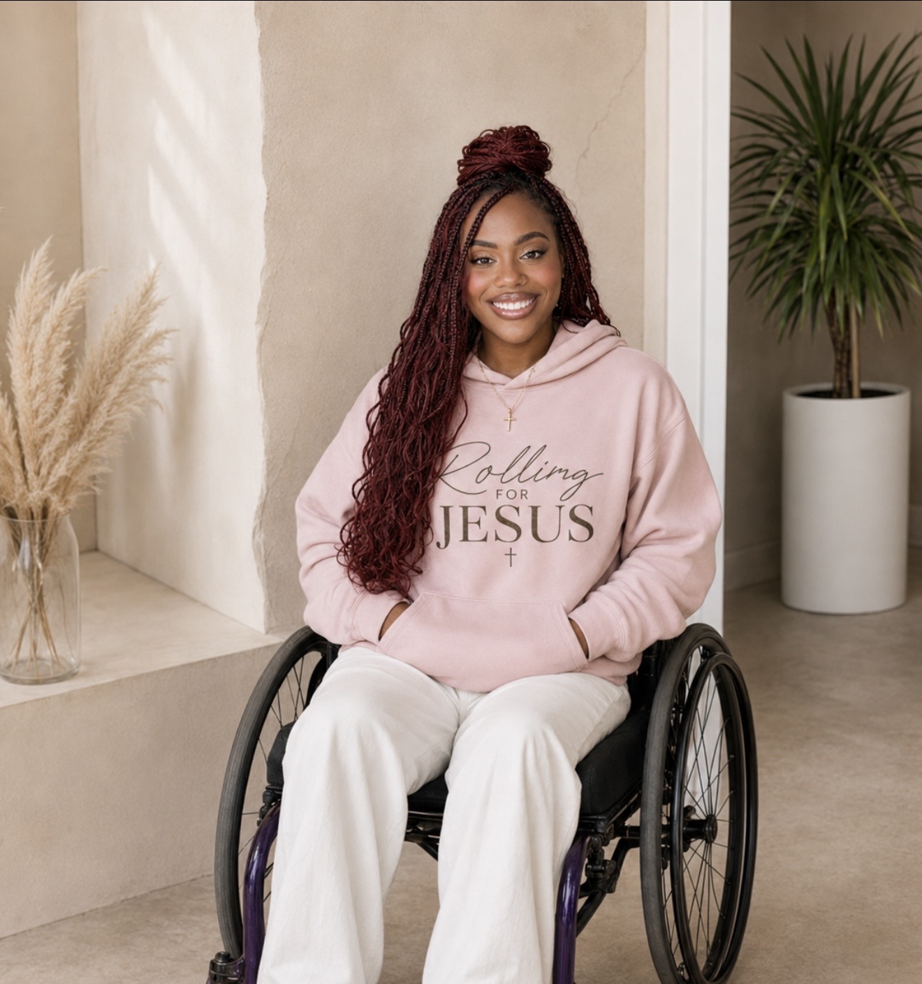 Smiling woman in a pink hoodie and white pants sitting in a wheelchair indoors, with plants in the background.