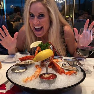Woman with blonde hair smiling and raising hands at a seafood platter with lobster, shrimp, lemon, and sauces, in a restaurant.