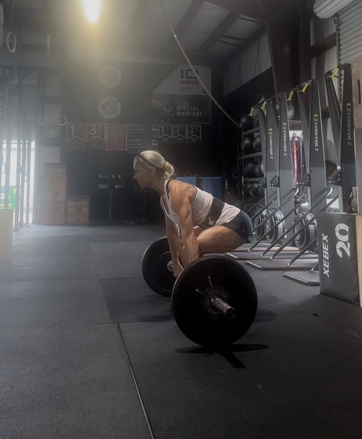 Woman lifting a barbell in a gym with equipment and chalkboard in background.