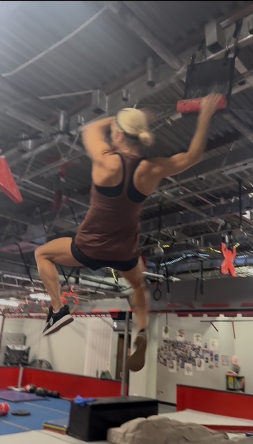 Person climbing a rope in an indoor gym, wearing a tank top and shorts, with gym equipment visible in the background.