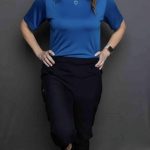 Woman standing with hands on hips, smiling, wearing a blue shirt and black pants, against a gray background.