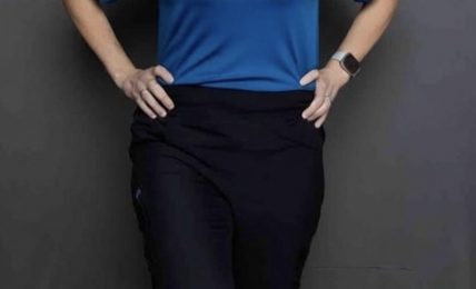Woman standing with hands on hips, smiling, wearing a blue shirt and black pants, against a gray background.