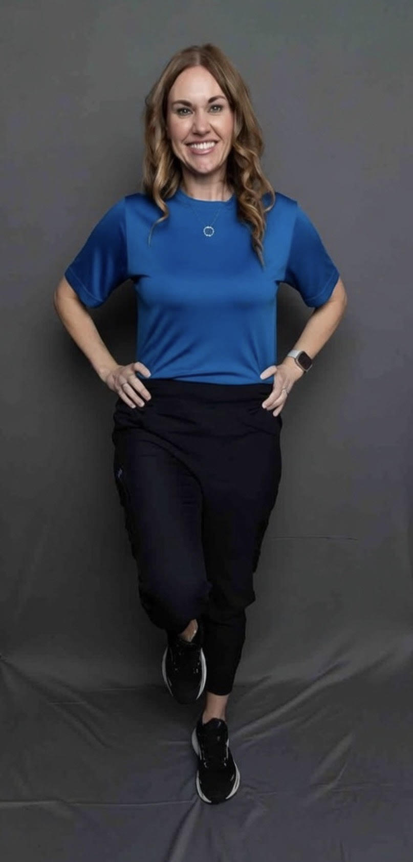 Woman standing with hands on hips, smiling, wearing a blue shirt and black pants, against a gray background.