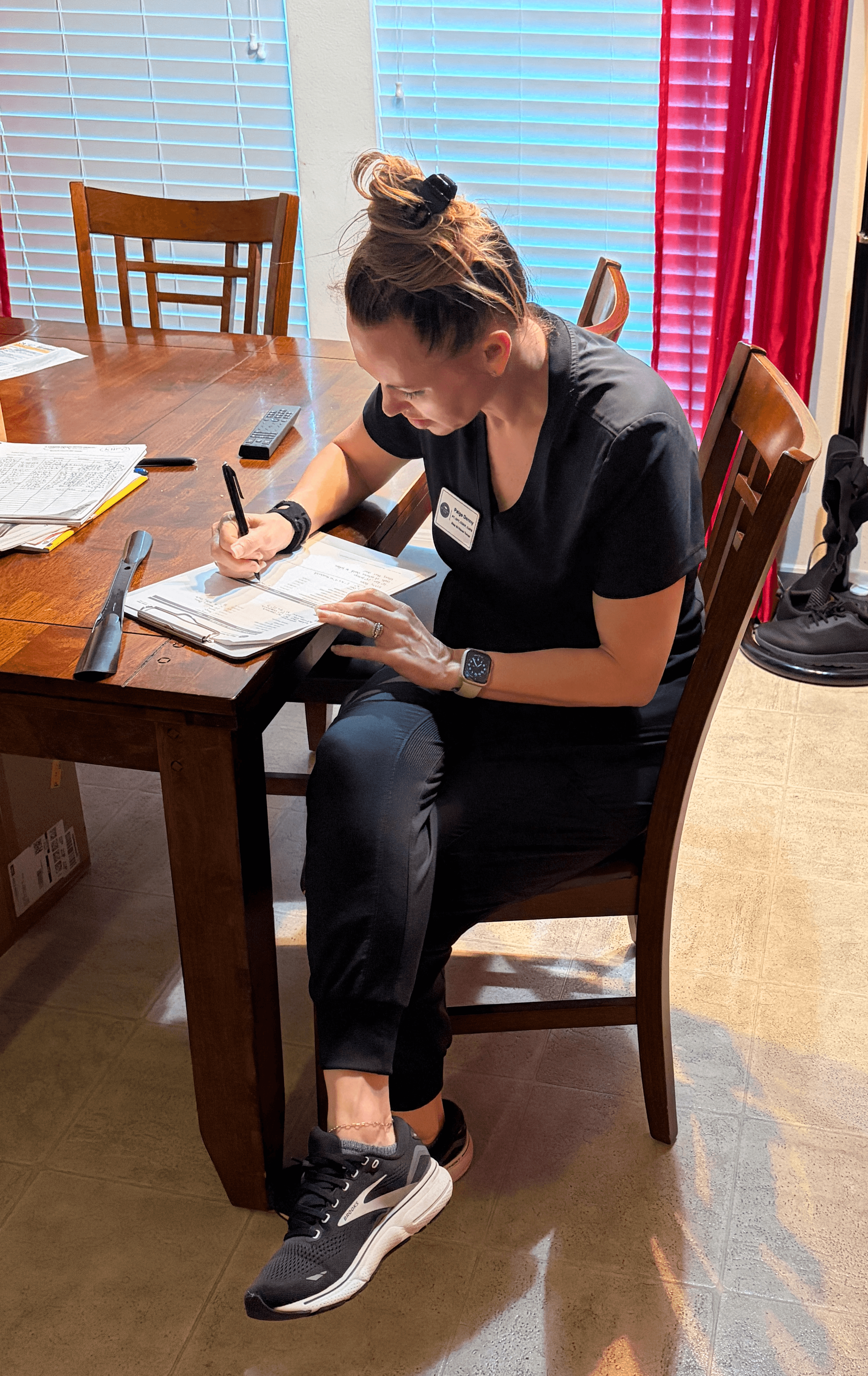 Woman sitting at a table writing in a notebook, with books and a pen nearby, in a room with colorful curtains.