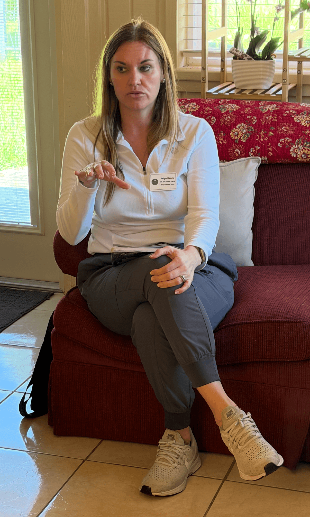 Woman sitting on a red couch, gesturing with her right hand, in a room with a window and plants behind her.