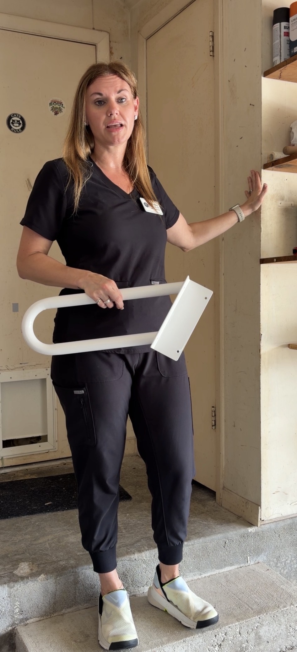 Woman holding a white object, standing in a room with shelves and a door, wearing dark scrubs and sneakers.