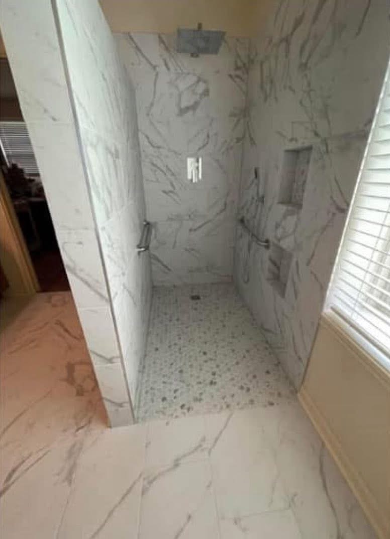 Small shower with marble walls and floor, two built-in shelves, and a window with blinds nearby.