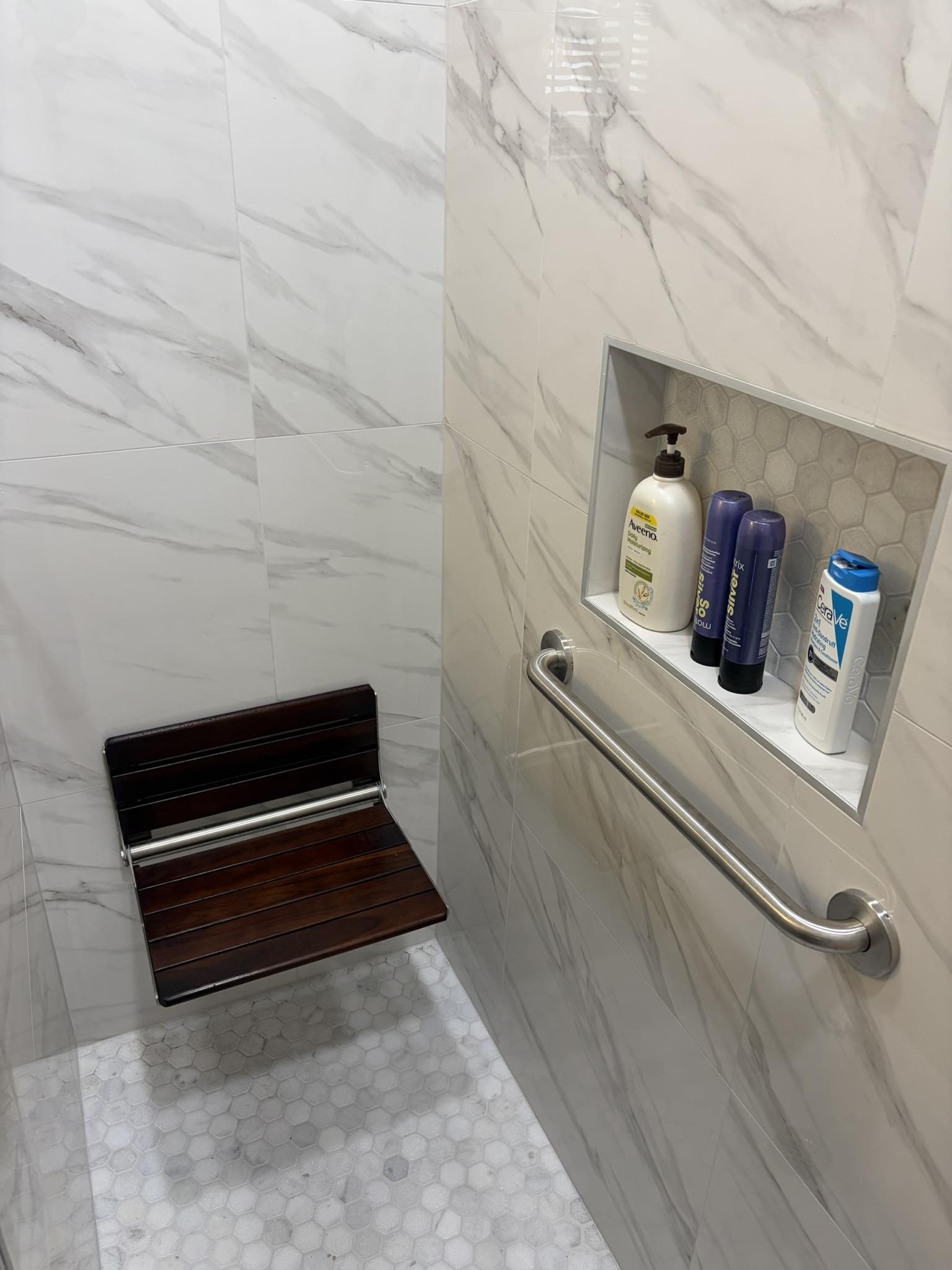 Bathroom corner with marble walls, a small wooden seat, and a built-in shelf with toiletries.