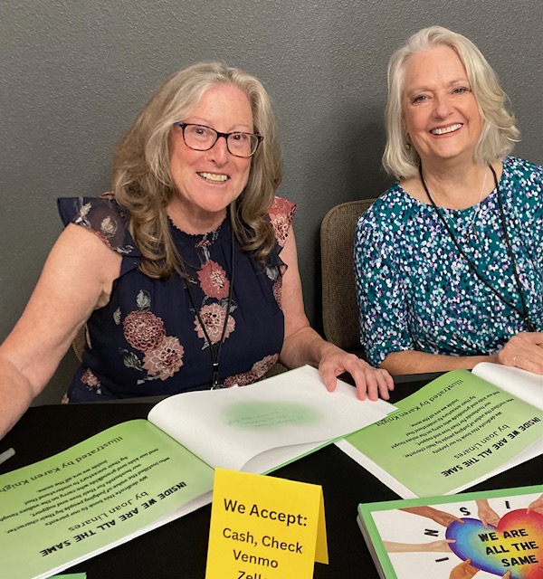Two women sitting at a table with open books and papers, smiling, in a gray room.