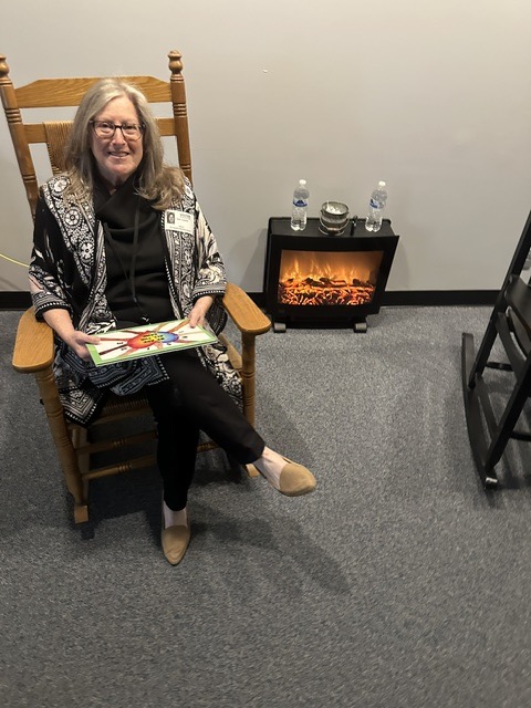 Woman sitting in a wooden chair holding a tray, with a small electric fireplace and water bottles behind her, in a room with gray carpet.