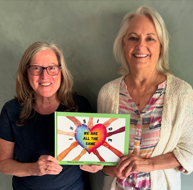 Two women holding a colorful sign with a heart and hands, smiling, against a green wall.