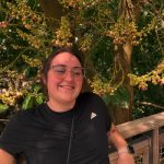 Smiling woman with glasses and black athletic shirt standing outdoors near a wooden railing, surrounded by yellow flowering tree branches.