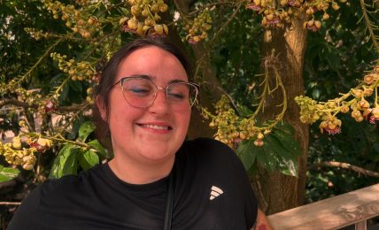 Smiling woman with glasses and black athletic shirt standing outdoors near a wooden railing, surrounded by yellow flowering tree branches.