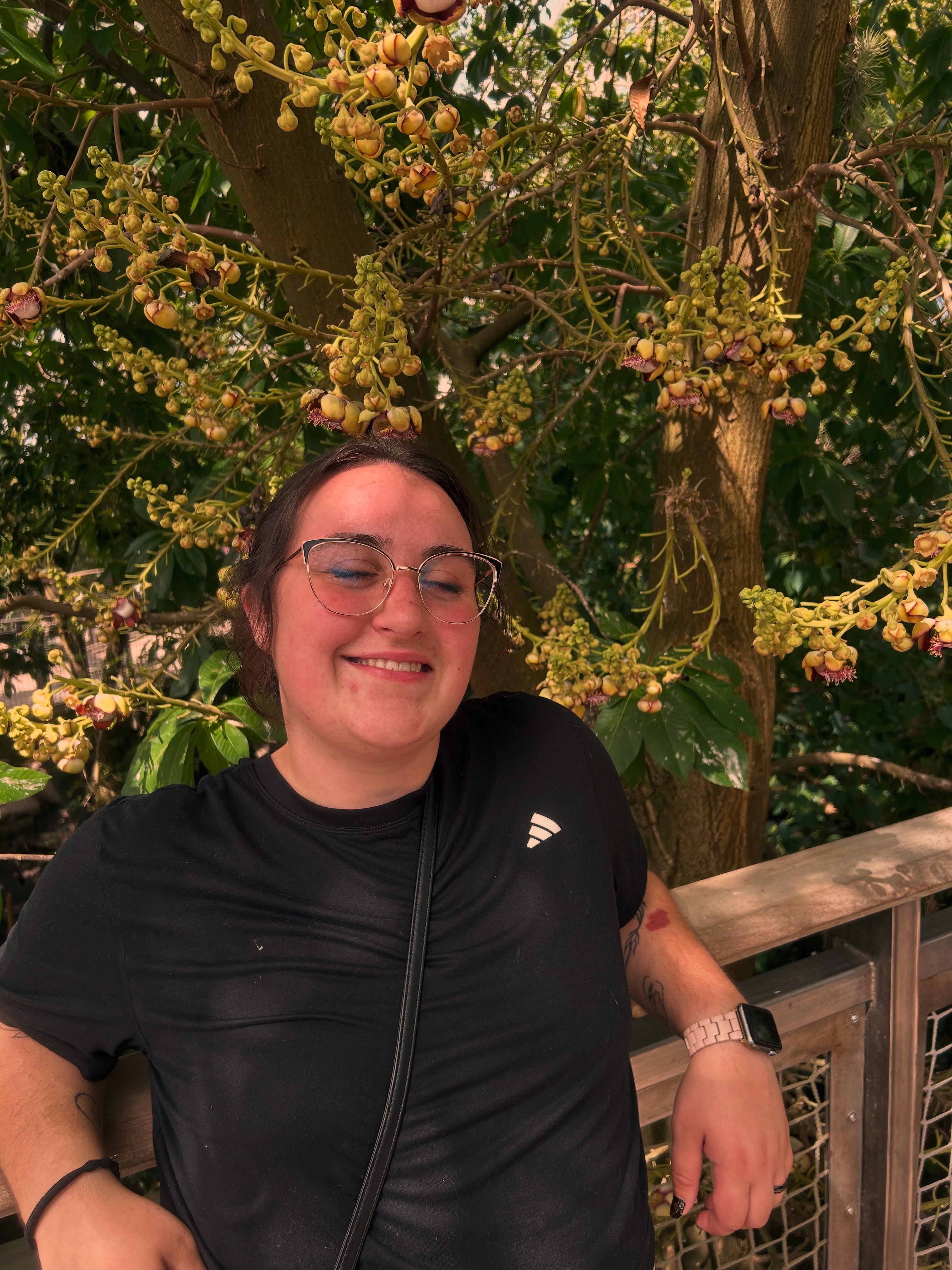 Smiling woman with glasses and black athletic shirt standing outdoors near a wooden railing, surrounded by yellow flowering tree branches.