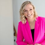 Smiling woman with blonde wavy hair wearing a bright pink blazer, standing indoors against a light background.