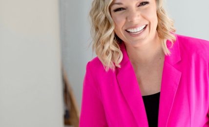 Smiling woman with blonde wavy hair wearing a bright pink blazer, standing indoors against a light background.