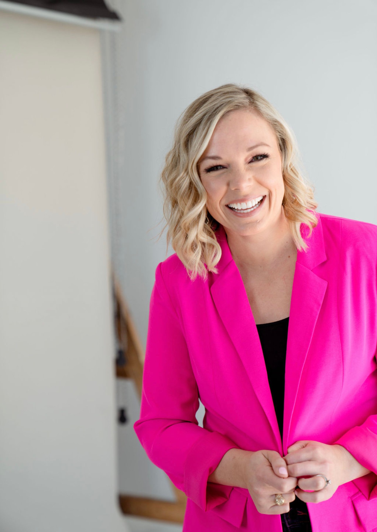 Smiling woman with blonde wavy hair wearing a bright pink blazer, standing indoors against a light background.