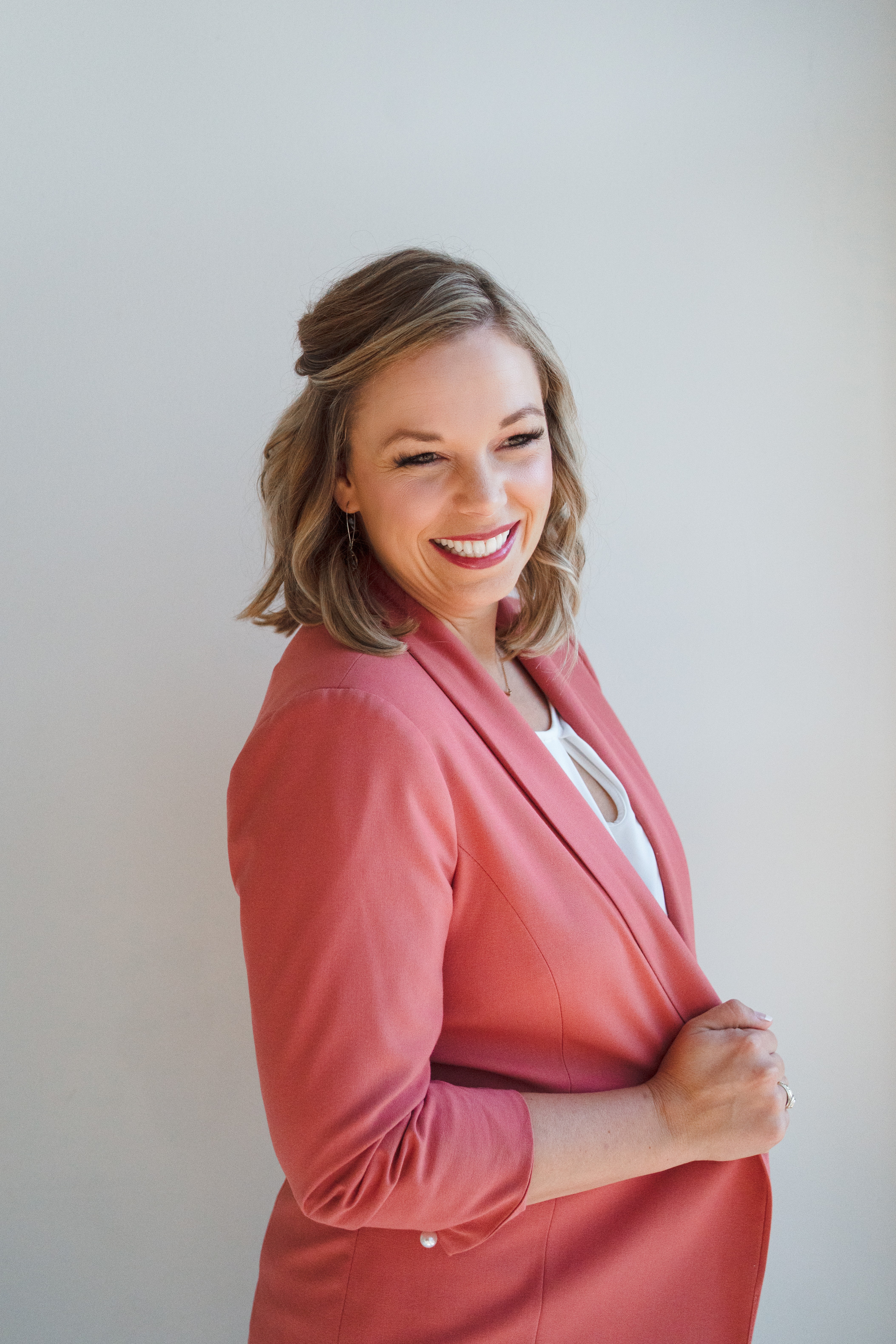 Smiling woman with shoulder-length hair wearing a pink blazer and white top, standing against a plain background.