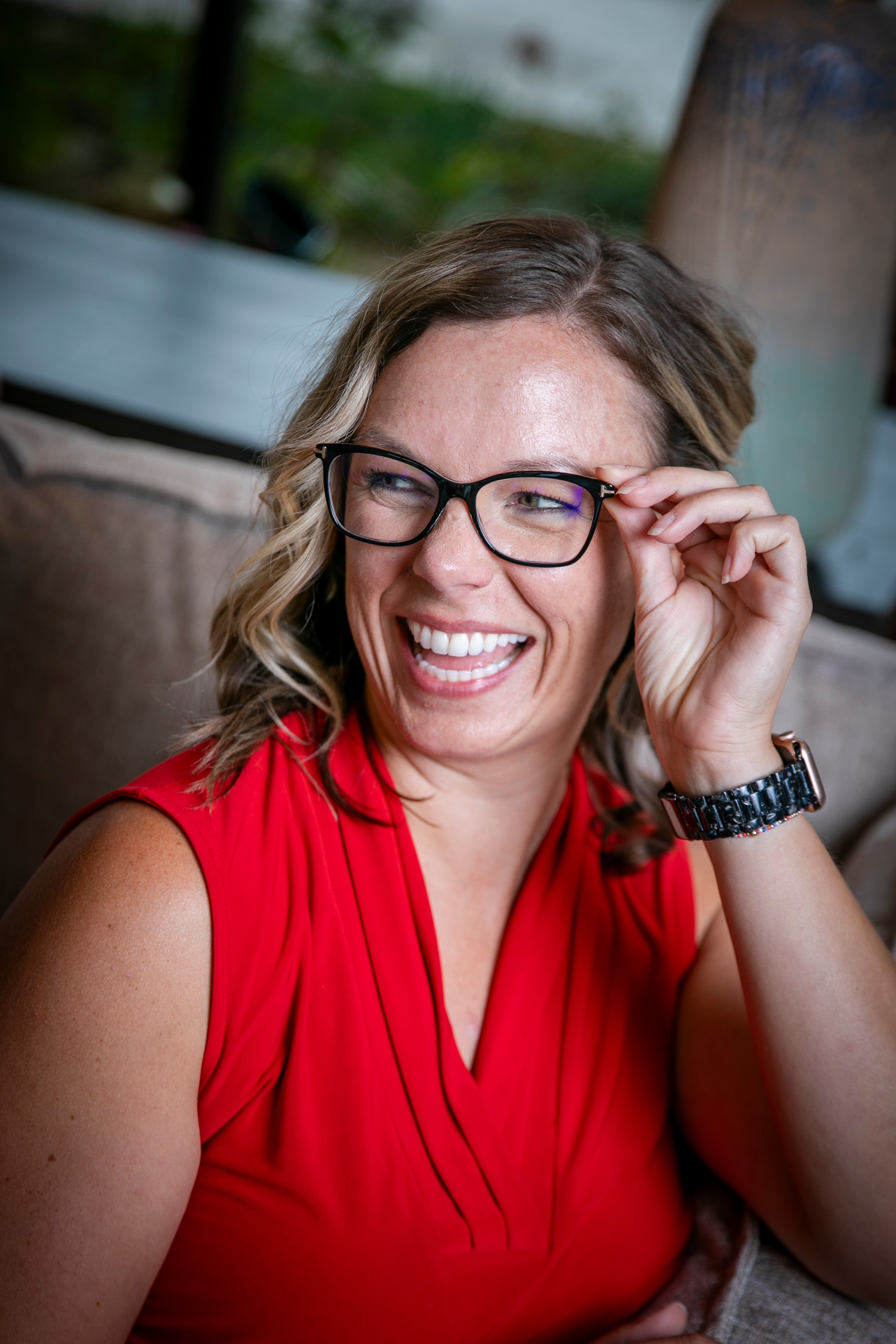 Woman with glasses smiling and adjusting her glasses, wearing a red sleeveless top, sitting indoors.