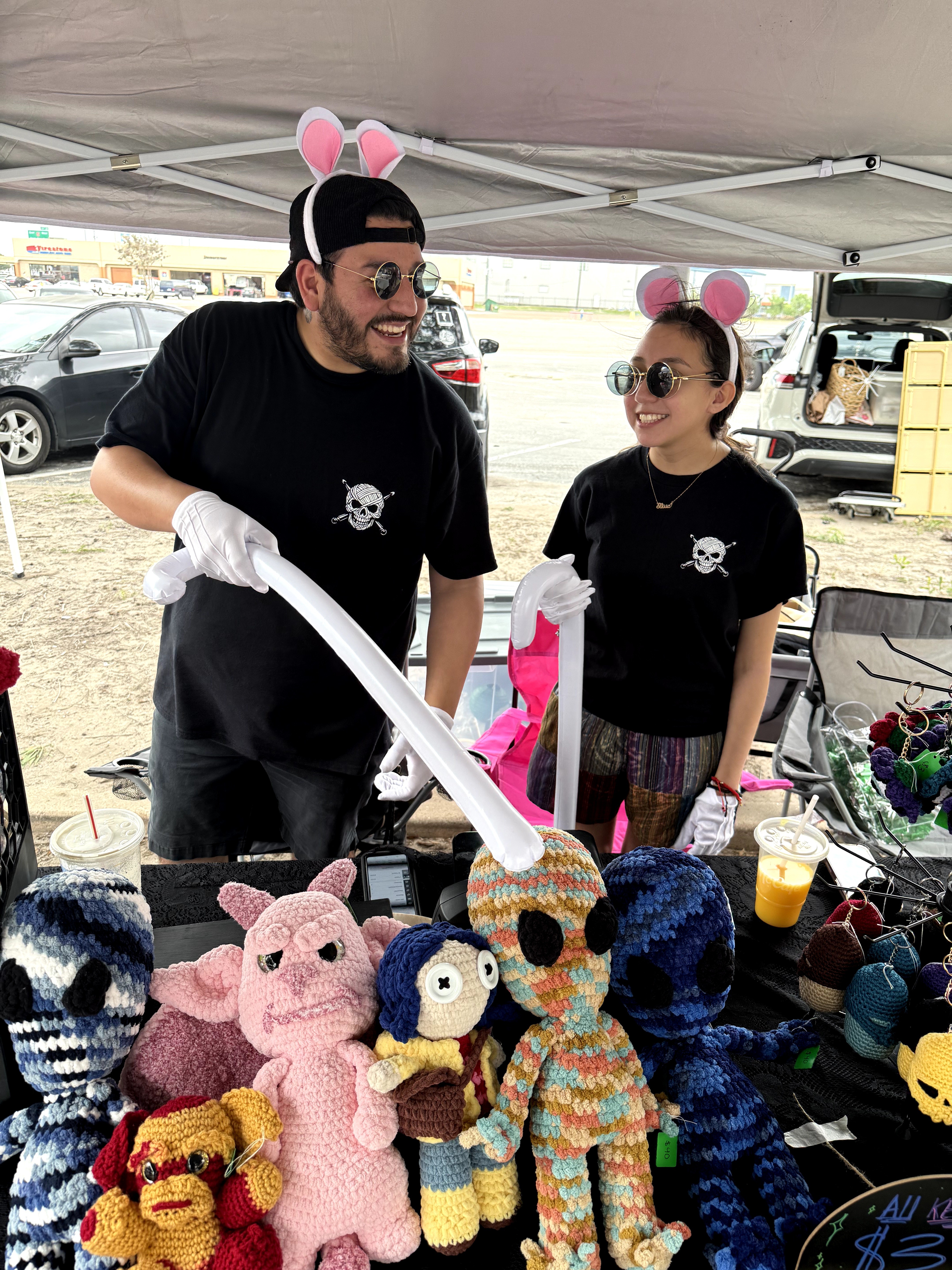 Two people wearing pink bunny ears and sunglasses at a craft booth with knitted animal toys, including a pig, penguin, and dog.