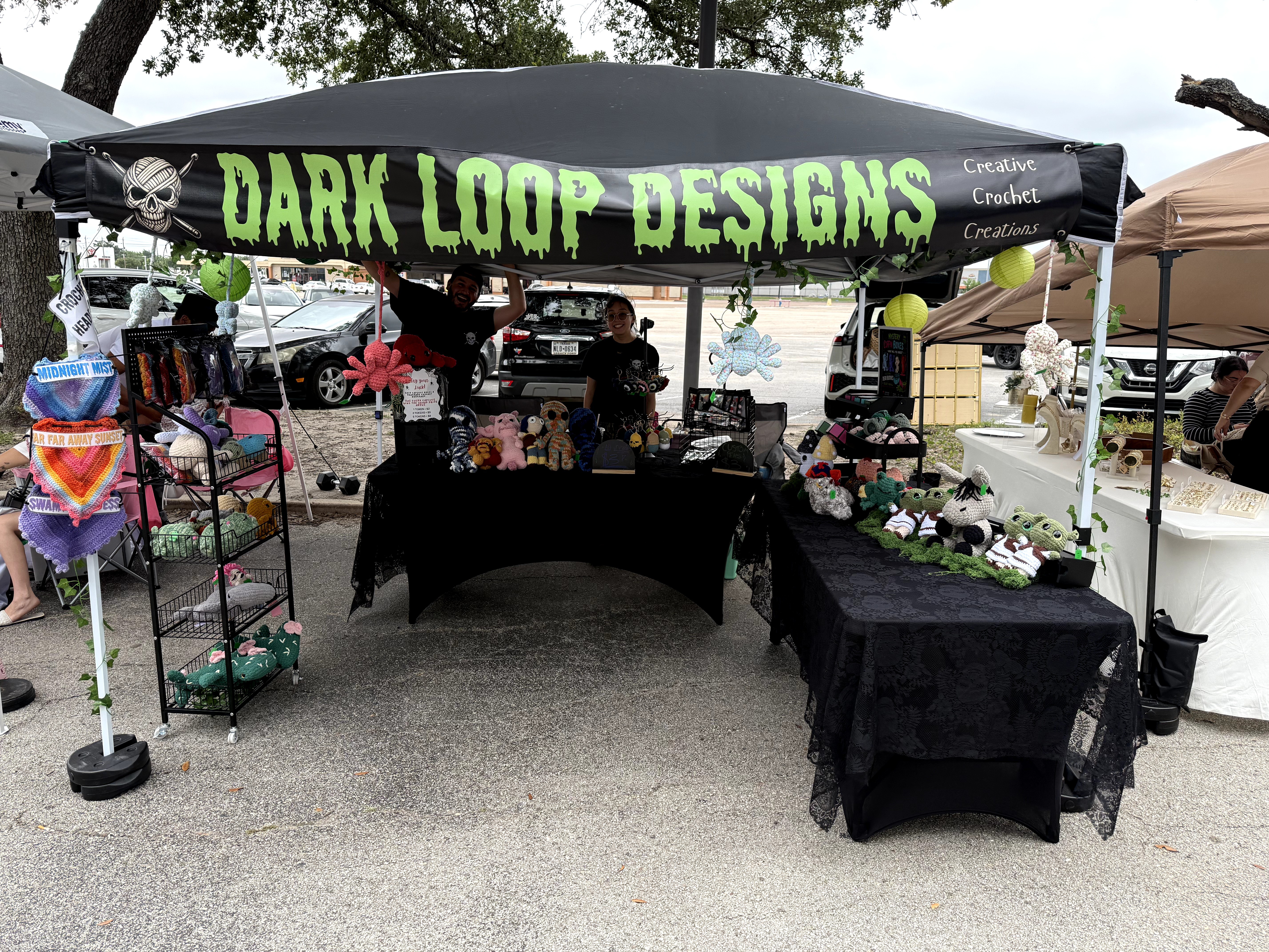 Outdoor market stall with a black canopy labeled 'Dark Loop Designs,' displaying jewelry and crafts on tables and shelves.
