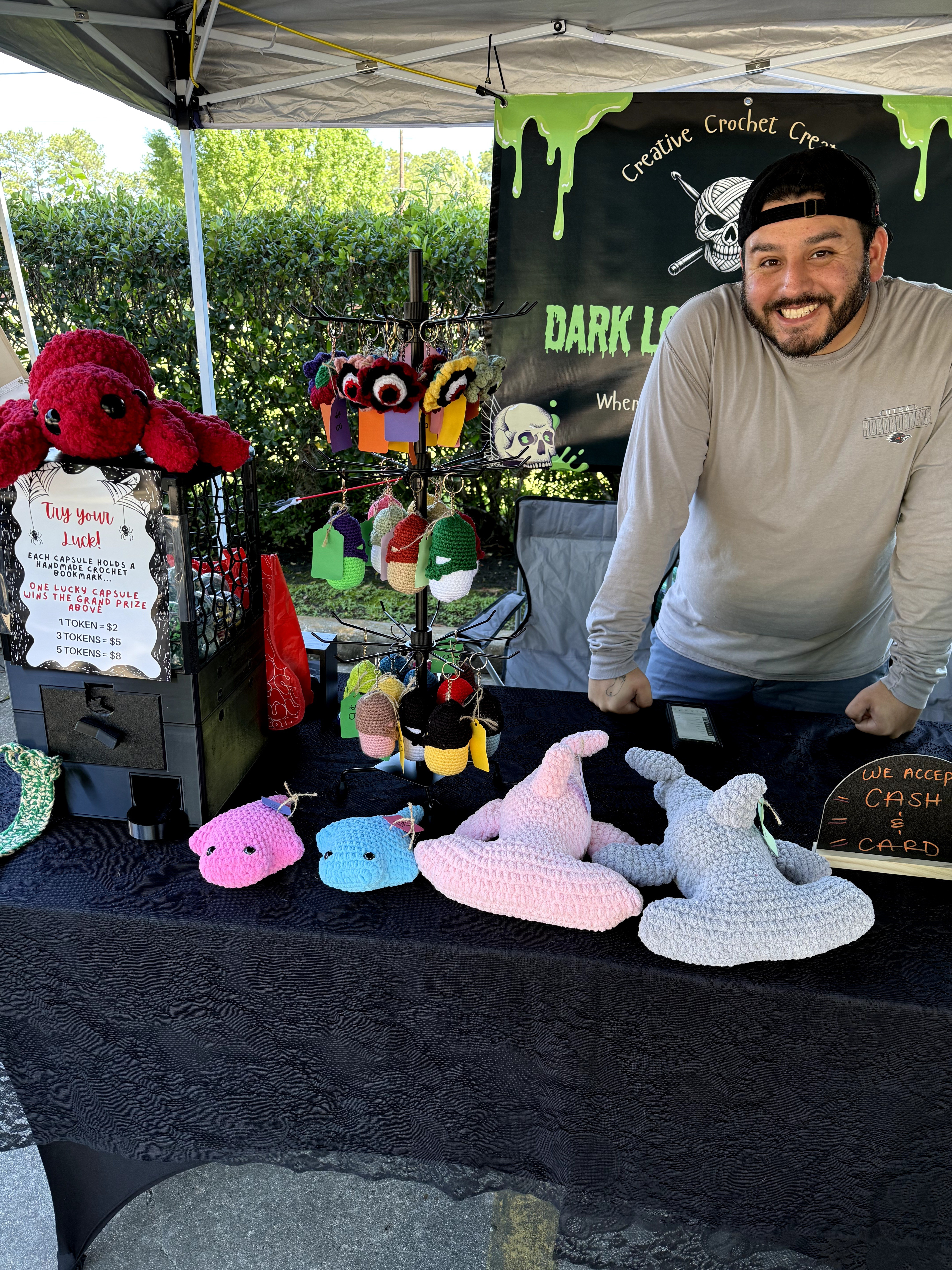 Man smiling at craft crochet booth with colorful plush toys and a dark-themed banner, outdoors with greenery in background.