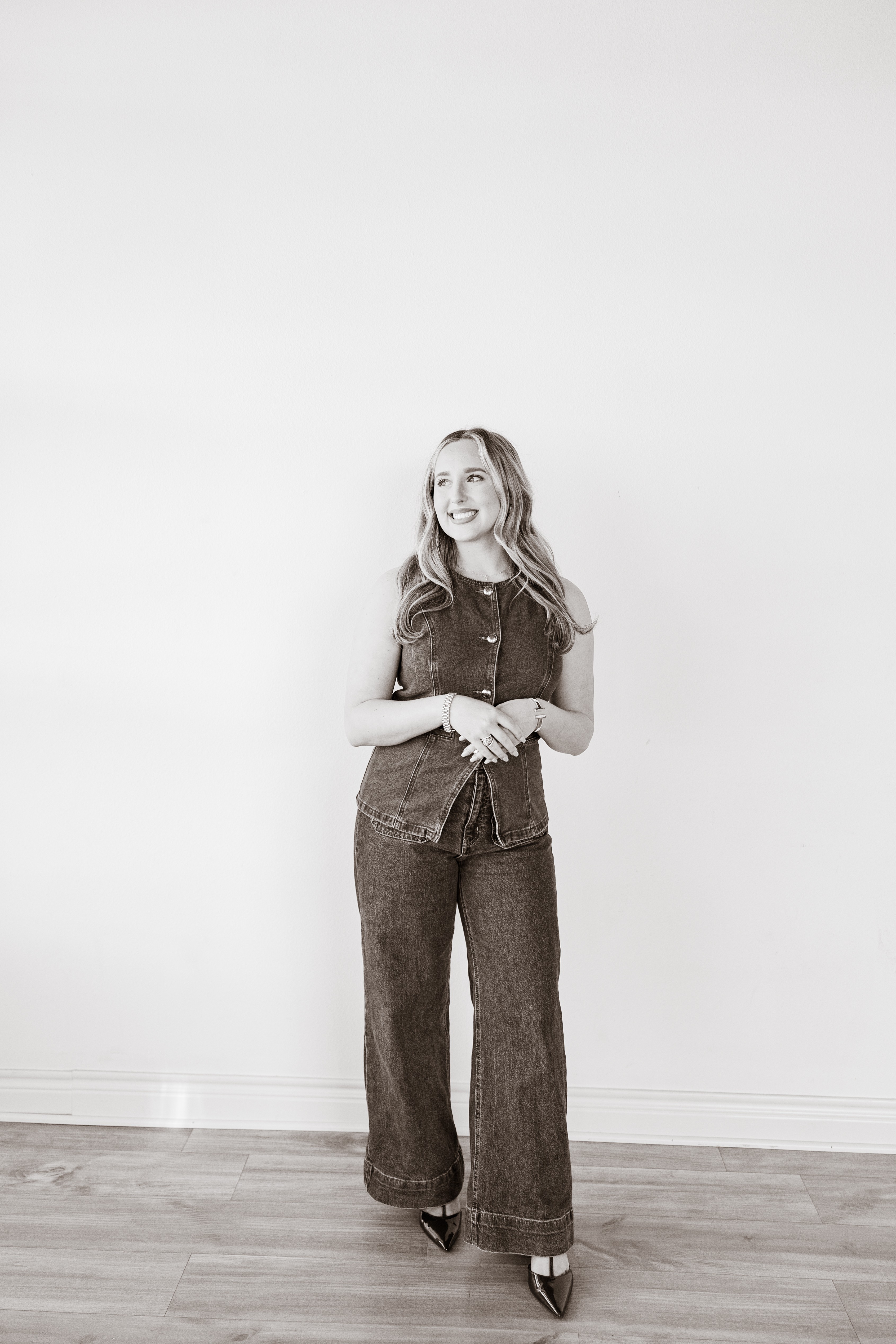 Young woman with long hair standing against a plain white wall, smiling, wearing a sleeveless top and wide-leg jeans, hands clasped.