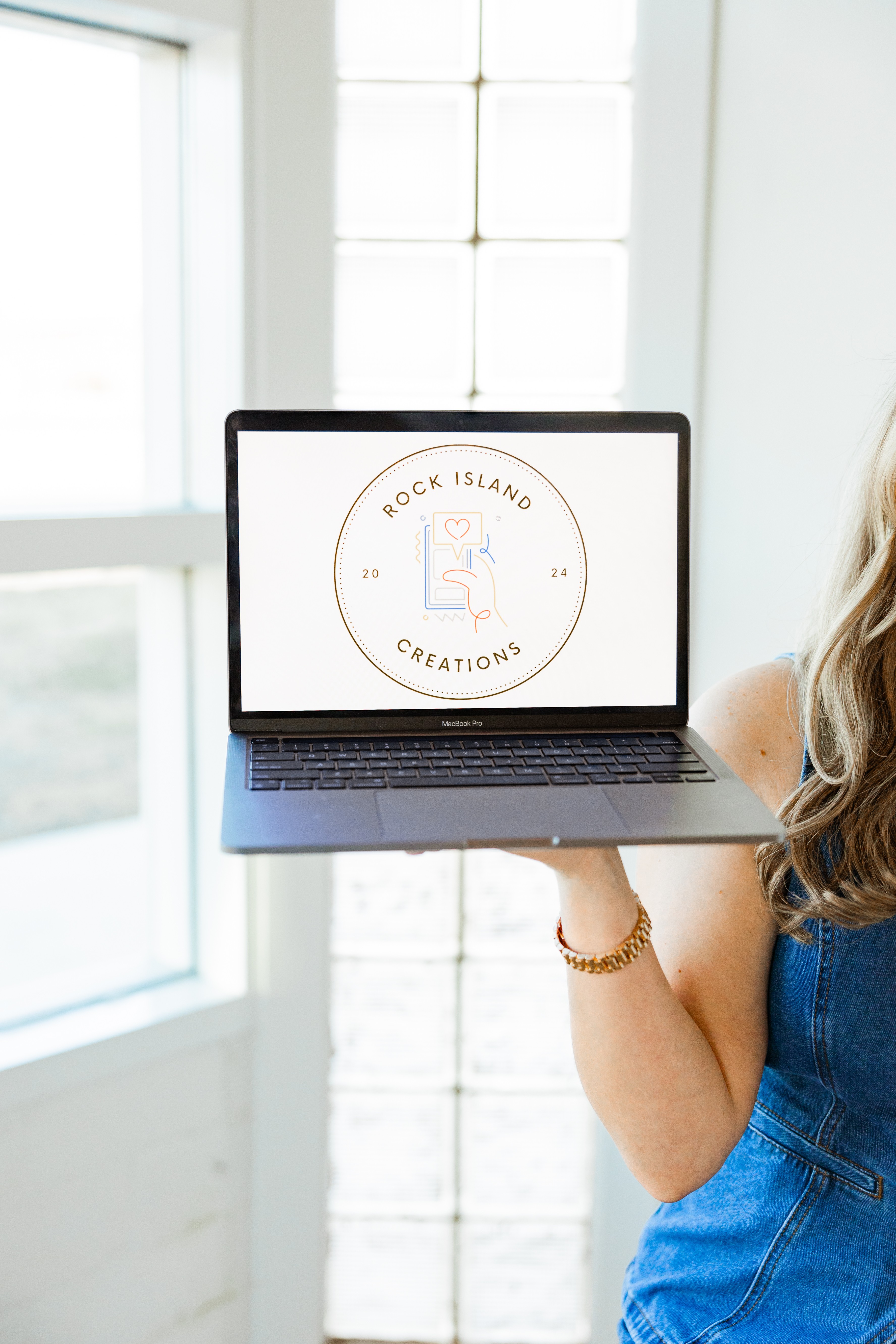 Person holding a laptop displaying a logo with a hand and text, in front of a window with white curtains