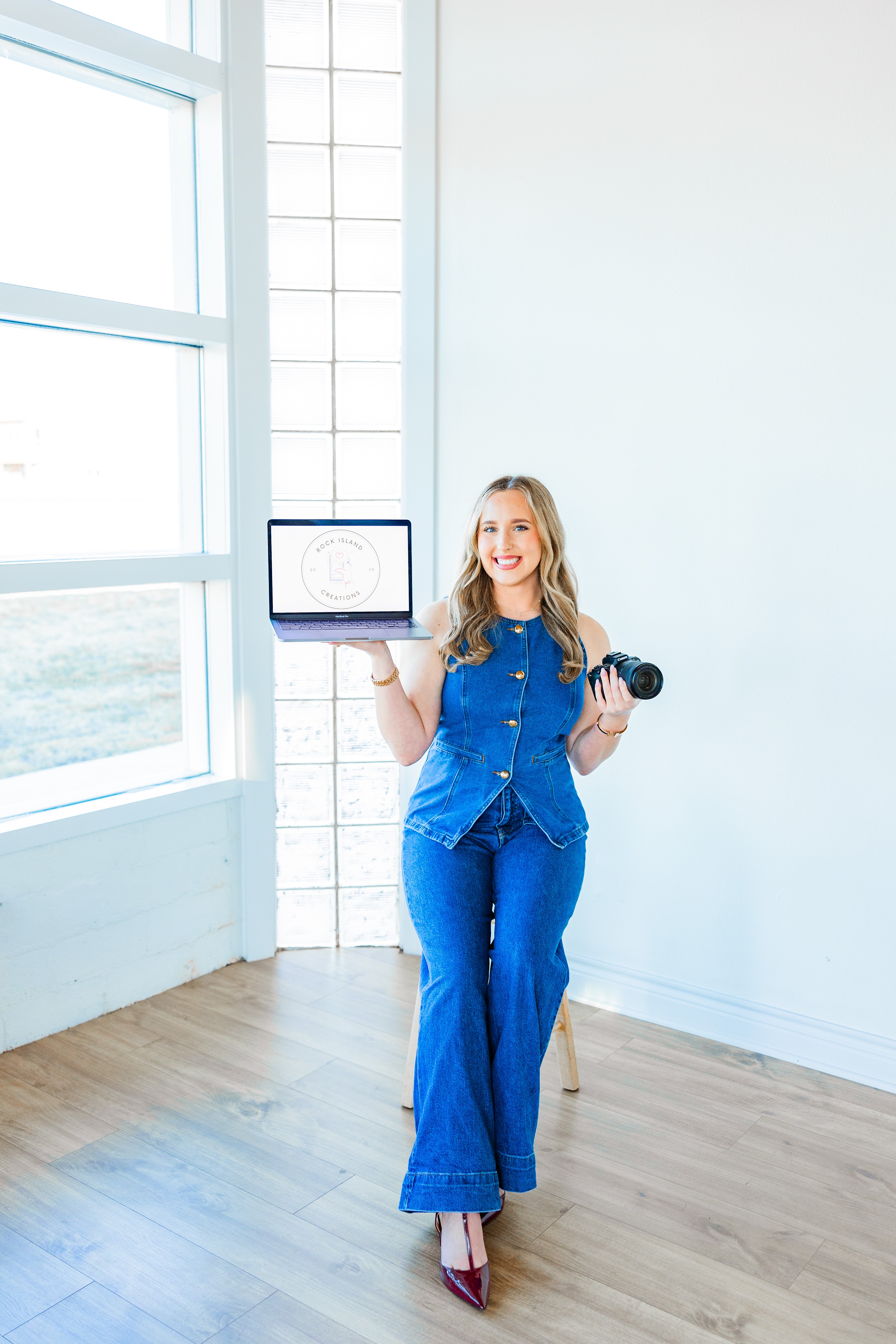 Woman with long blonde hair holding a tablet and camera, standing in a bright room with large windows.