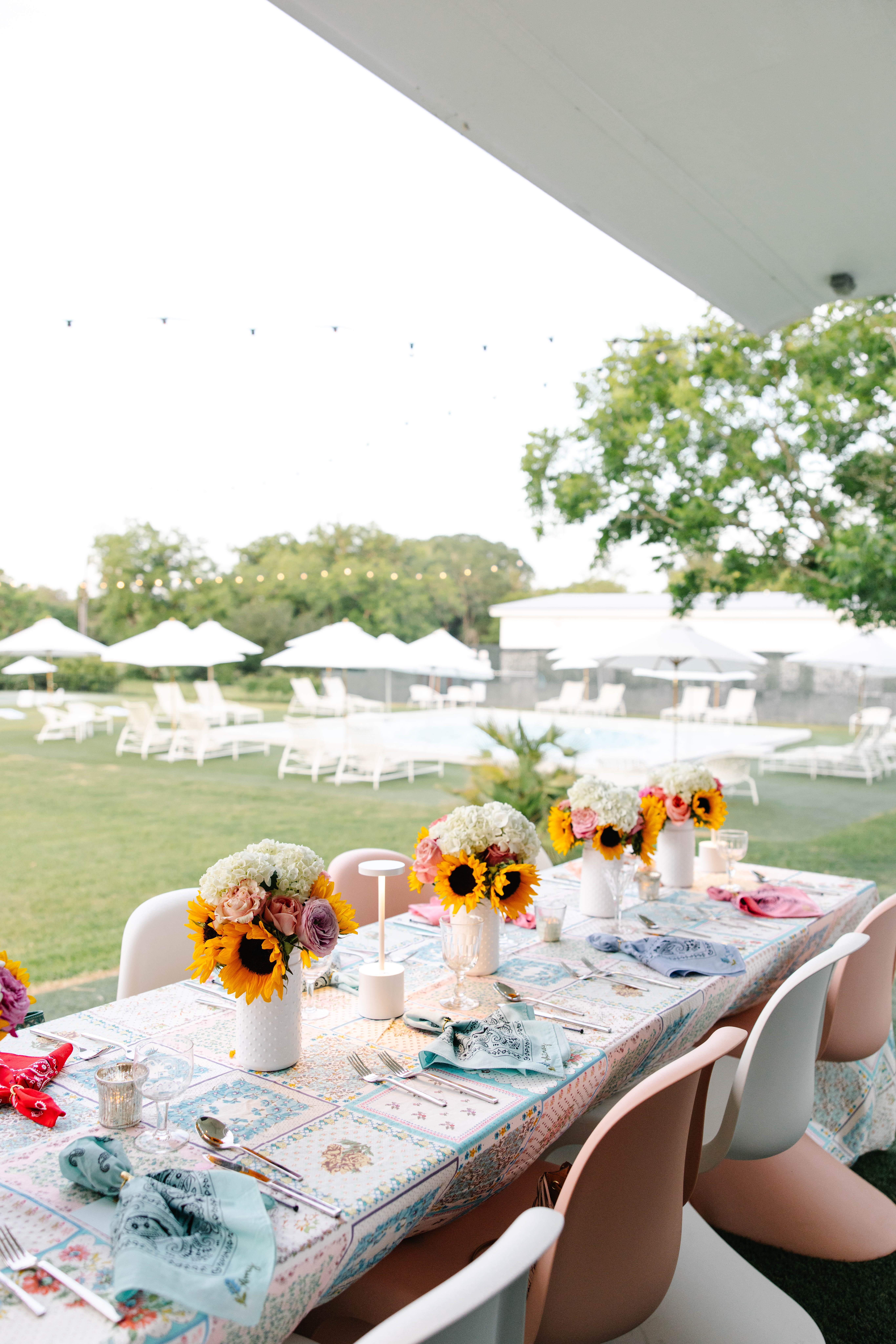 Table set outdoors with floral arrangements, tableware, and chairs, under a large canopy with trees and tents in the background.