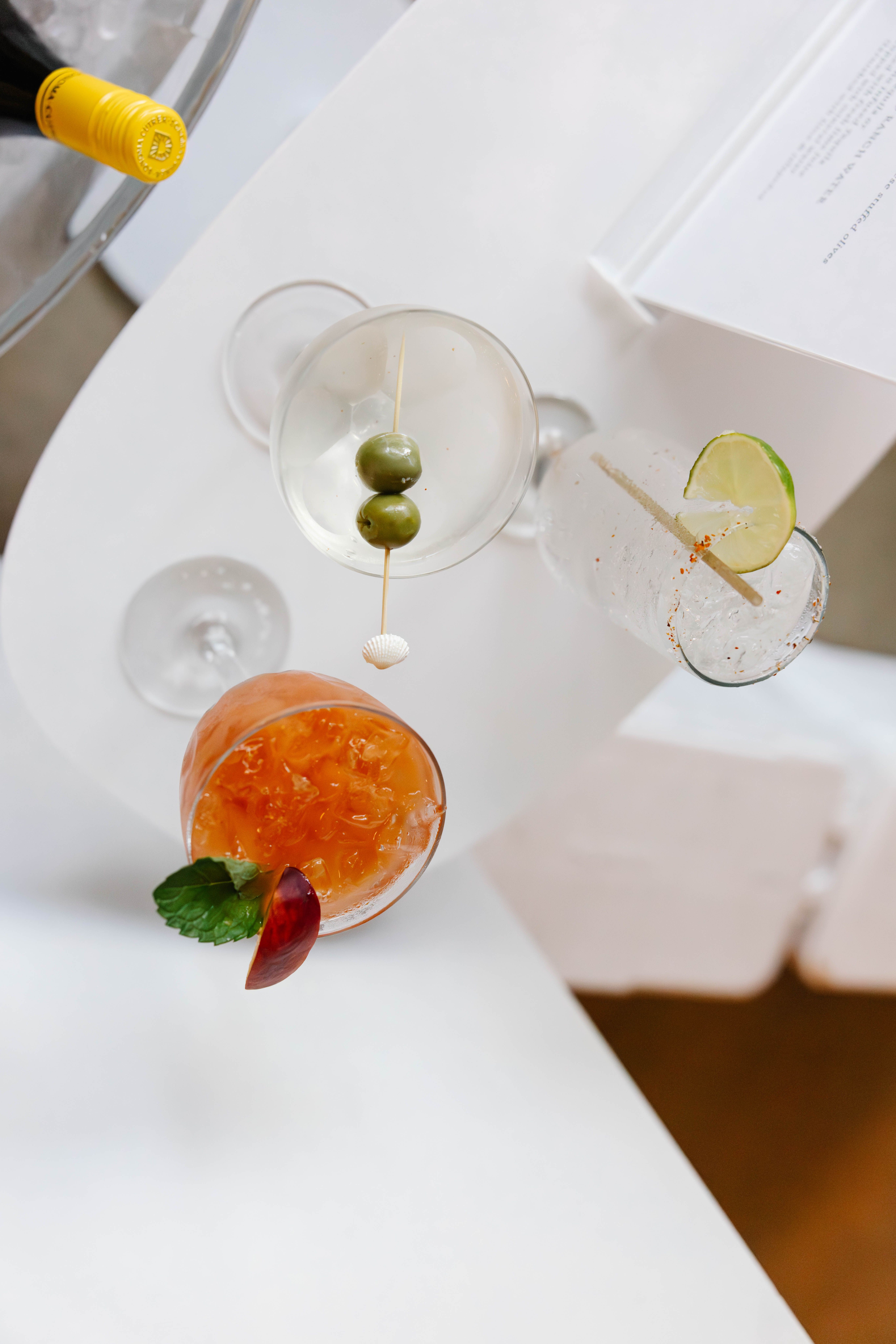 Cocktail glass with orange drink, garnished with a red fruit slice and mint leaf, on a white surface.