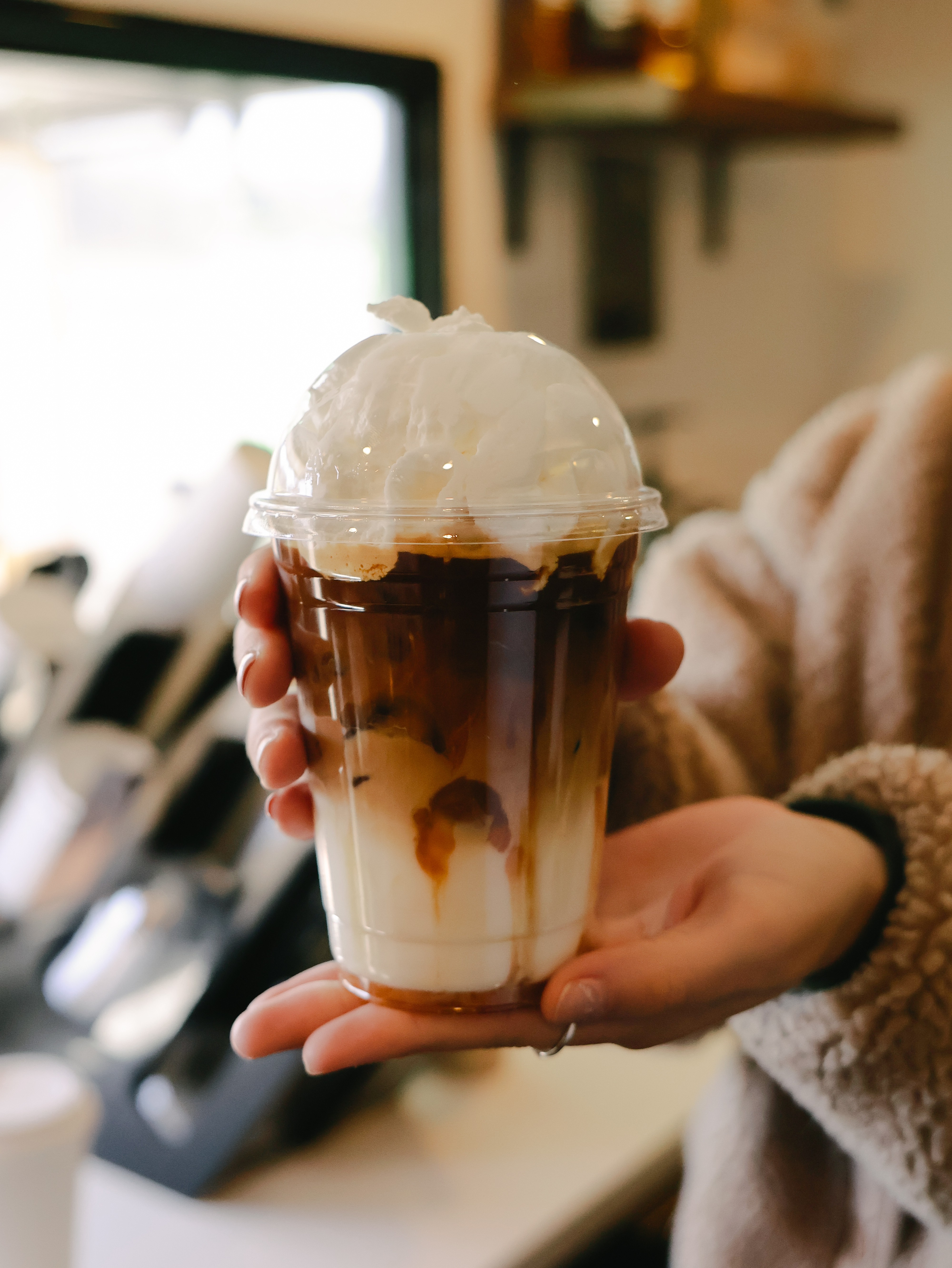 Person holding a layered iced coffee with whipped cream, inside a cafe with a window and shelves in the background.