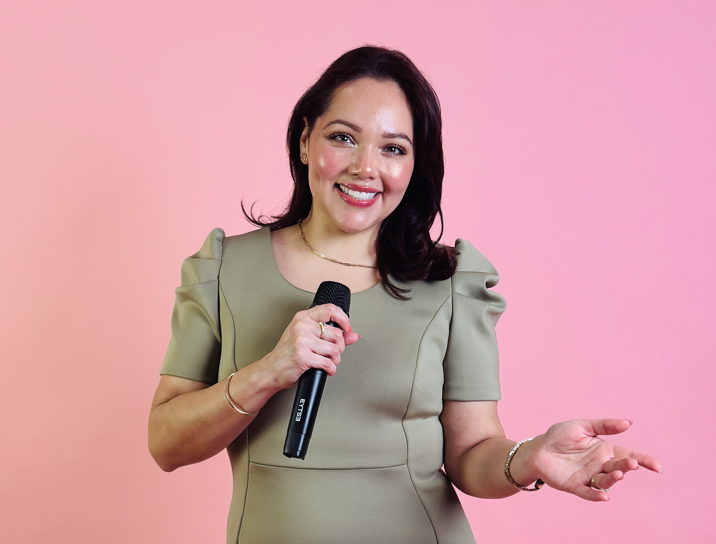 Woman smiling holding a microphone, gesturing with her other hand, against a pink background.
