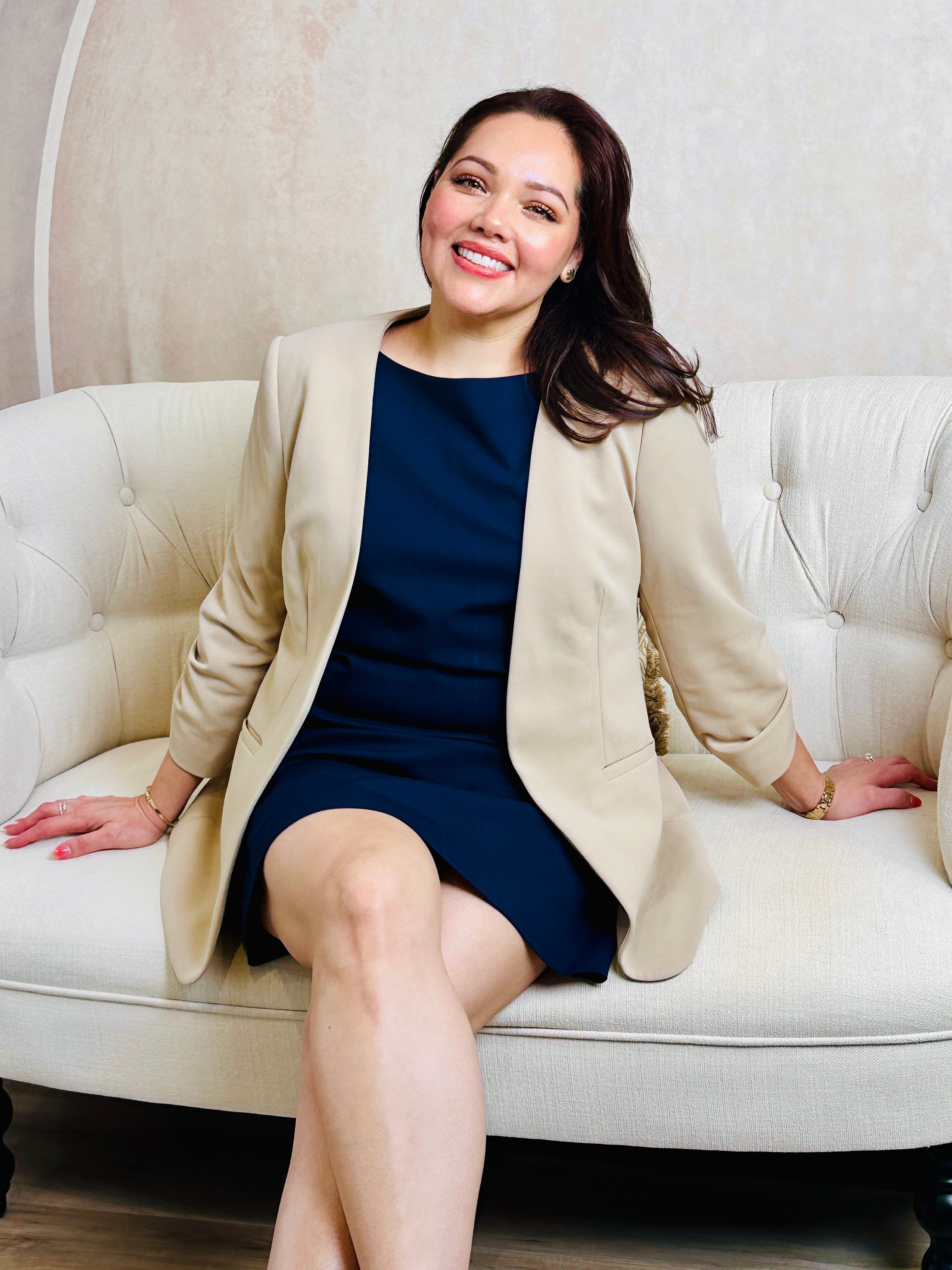 Woman sitting on a white sofa, smiling, wearing a navy dress and beige blazer, with hands resting on sofa.