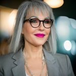 A woman with gray hair, glasses, and bright pink lipstick, wearing a gray blazer and pearl necklace, smiling.