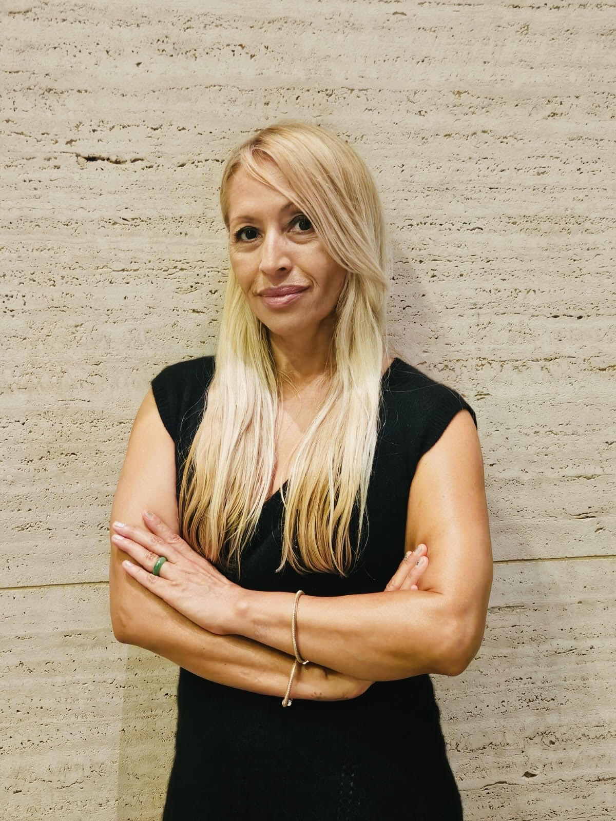 Woman with long blonde hair wearing a black sleeveless top, standing against a textured beige wall.
