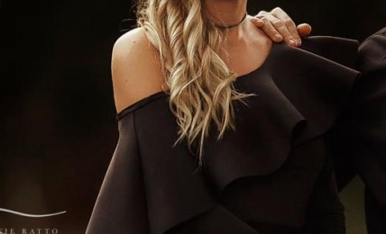 Woman with long wavy blonde hair wearing a black off-shoulder dress and choker necklace, standing indoors.