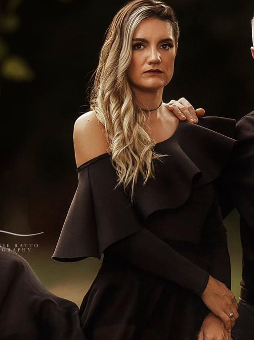 Woman with long wavy blonde hair wearing a black off-shoulder dress and choker necklace, standing indoors.