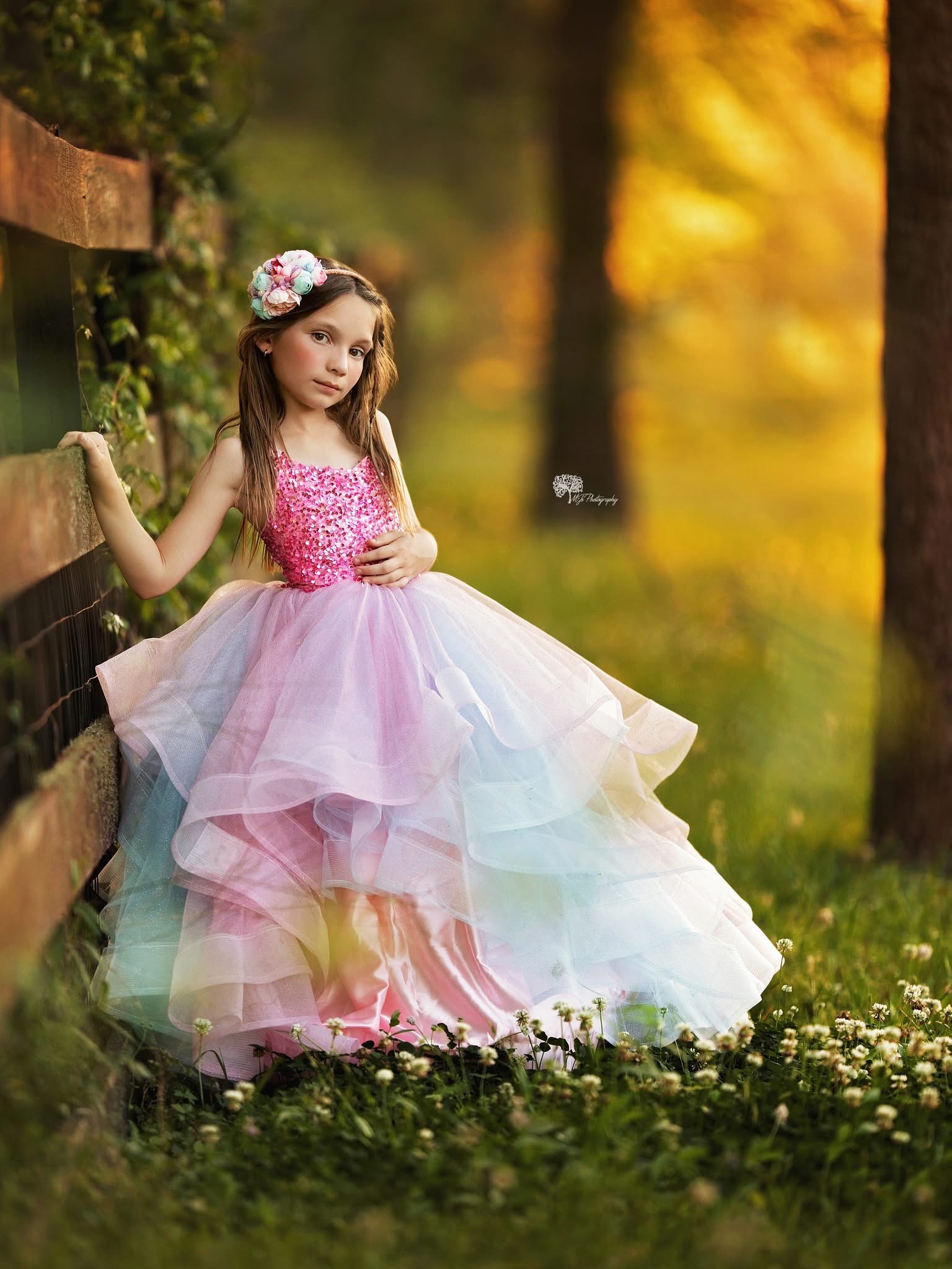 Young girl in a colorful, layered dress standing outdoors near a wooden fence, with trees and autumn foliage in background.