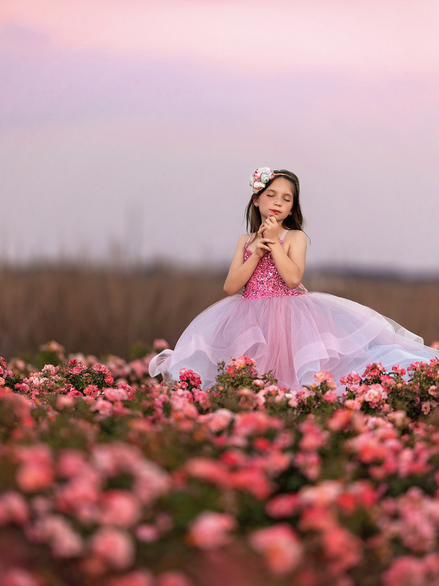 Young girl in a pink dress sitting among pink flowers outdoors, with a blurred background and a pink sky.
