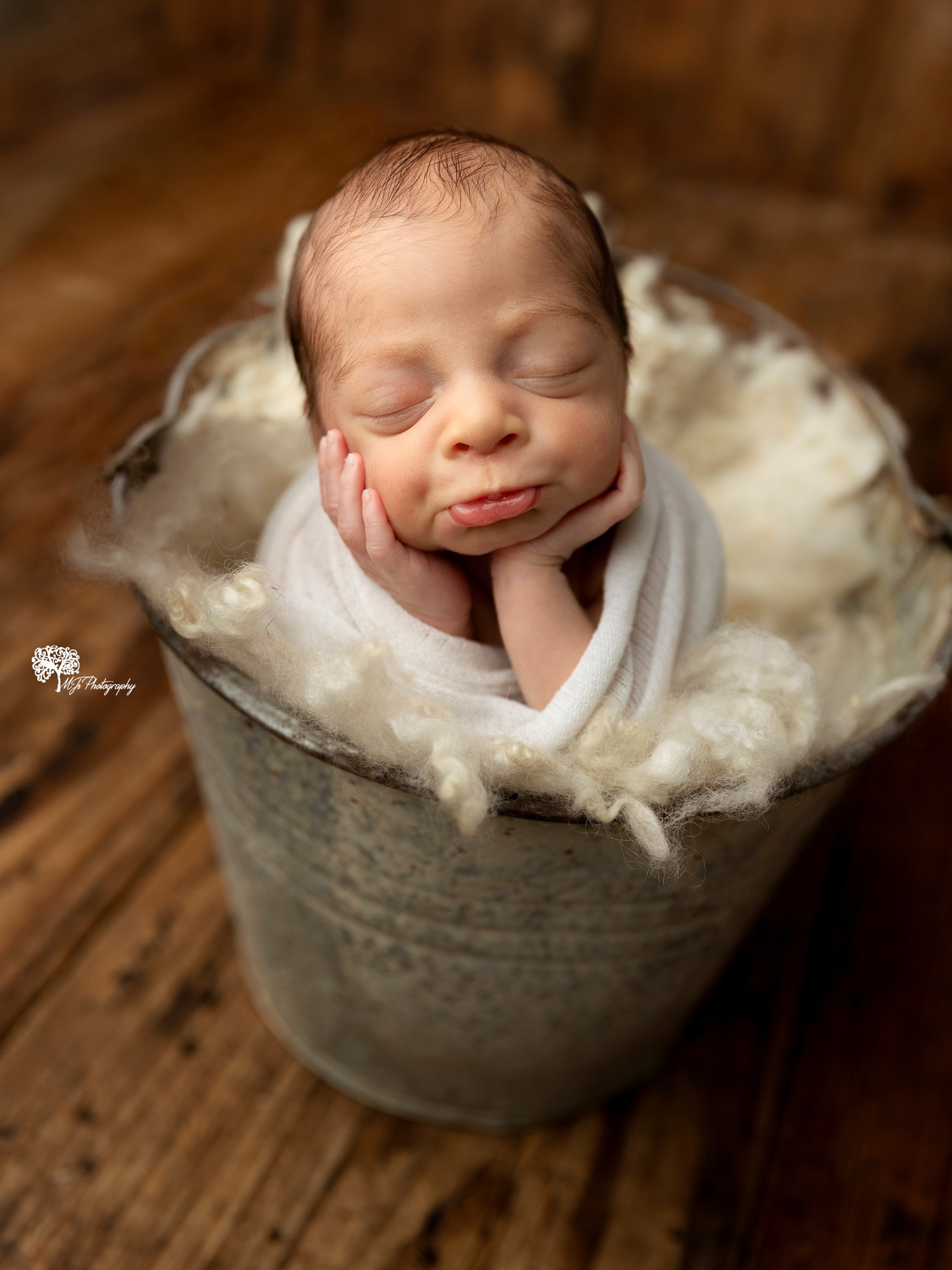 Newborn baby with closed eyes, resting chin on hands, wrapped in white cloth, in a metal bucket filled with soft material, on wooden floor.