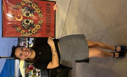 Woman standing next to a colorful poster with skulls and flowers, smiling, wearing a black top and checkered skirt, outdoors.