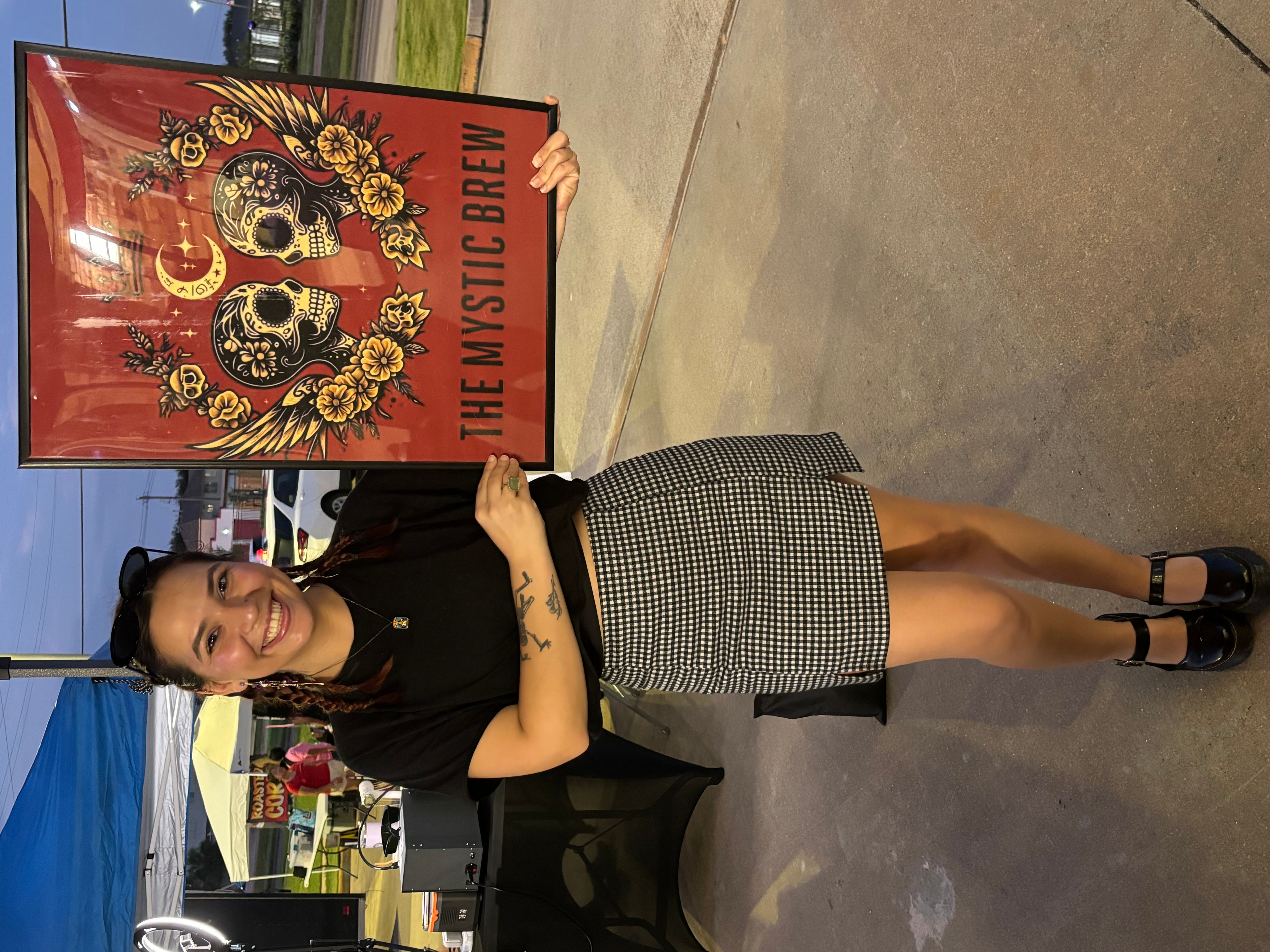 Woman standing next to a colorful poster with skulls and flowers, smiling, wearing a black top and checkered skirt, outdoors.